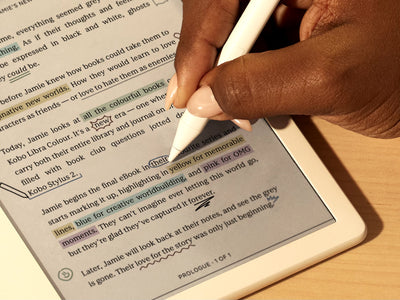 Een hand gebruikt de Kobo Stylus 2 om direct in een eBook te schrijven op een Kobo Libra Colour eReader op een vlak oppervlak. Op de pagina zien we kleurrijke aantekeningen en markeringen.