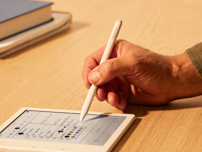 Een hand houdt een Kobo Stylus 2 boven een Kobo Libra Colour eReader op een vlak oppervlak. De punt van de stylus staat op het punt om een van de penkleuren te selecteren die in een optiemenu op het eReader-scherm worden weergegeven.