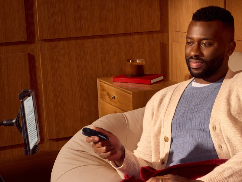 Un homme, confortablement assis, tient la Télécommande Kobo dans sa main droite, tout en regardant une liseuse Kobo sur un support flexible.